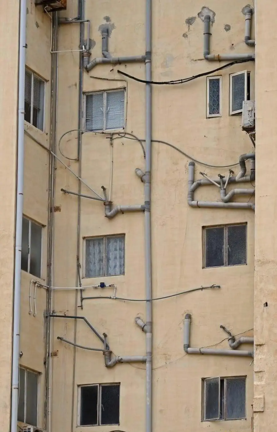 A beige building wall with several windows and a chaotic network of pipes and wires running in various directions, creating a tangled, disorganized appearance.