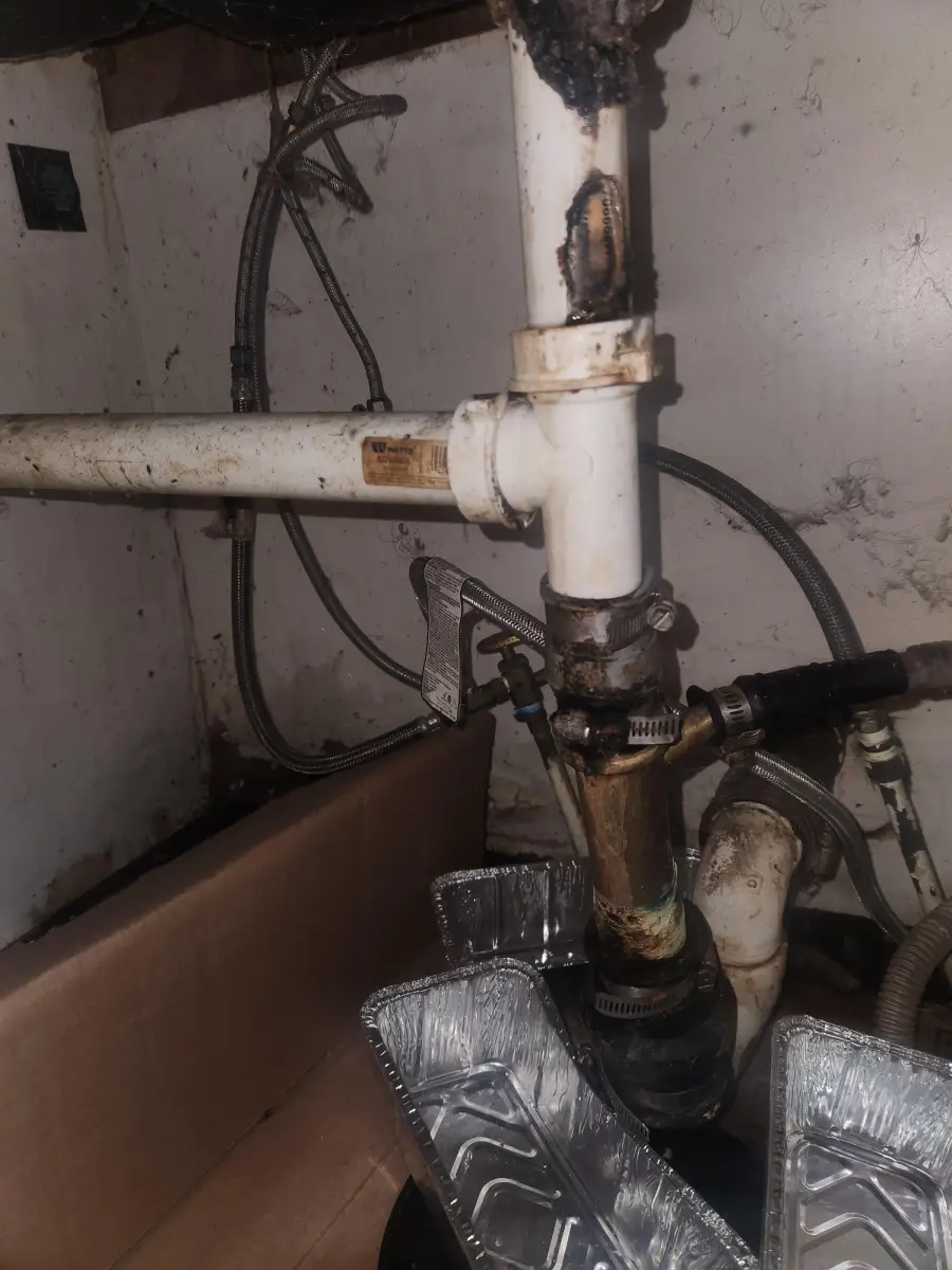 Dirty and corroded white pipes under a sink, with metal clamps and stains. Disposable foil trays and a cardboard box are placed below to catch leaks. Worn, stained wall in the background.