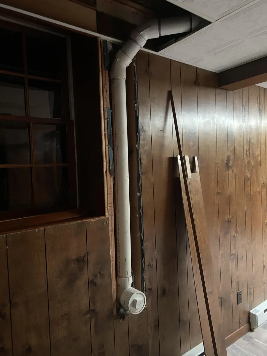 A basement with wood-paneled walls, a small window, vertical PVC piping, and two wooden boards leaning against the wall. The lighting is dim.