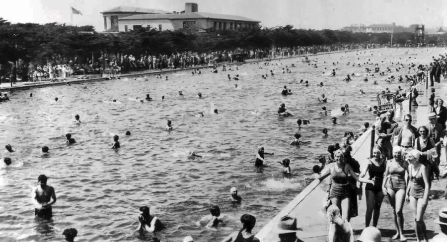 Black and white photo of a crowded outdoor pool with people swimming and standing along the edge; trees and a large building in the background; likely taken in the early 20th century.
