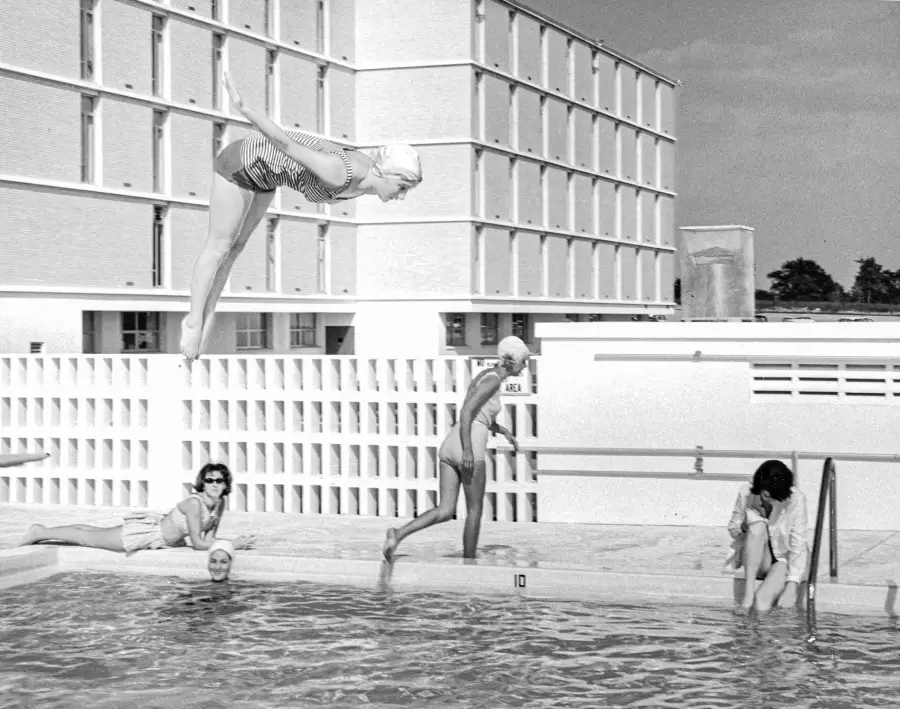 Four women are at a pool: one dives in midair, one walks on the pool edge, one sits on the poolside, and one sits on the pool steps. A large building stands in the background.