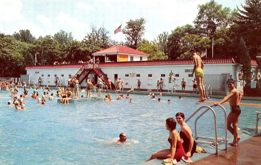 Large outdoor swimming pool crowded with people swimming and relaxing. Several people sit at the edge, while one person prepares to dive in. A building with an American flag is seen in the background.