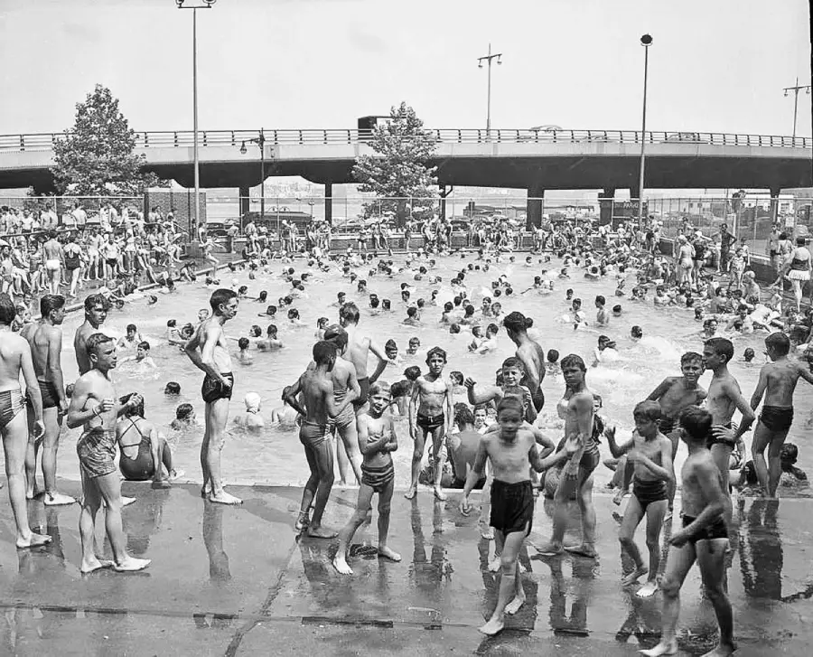 A crowded public swimming pool scene from the past, with many children and teenagers in swimwear, some standing on the poolside, others swimming or playing in the water, and an overpass in the background.