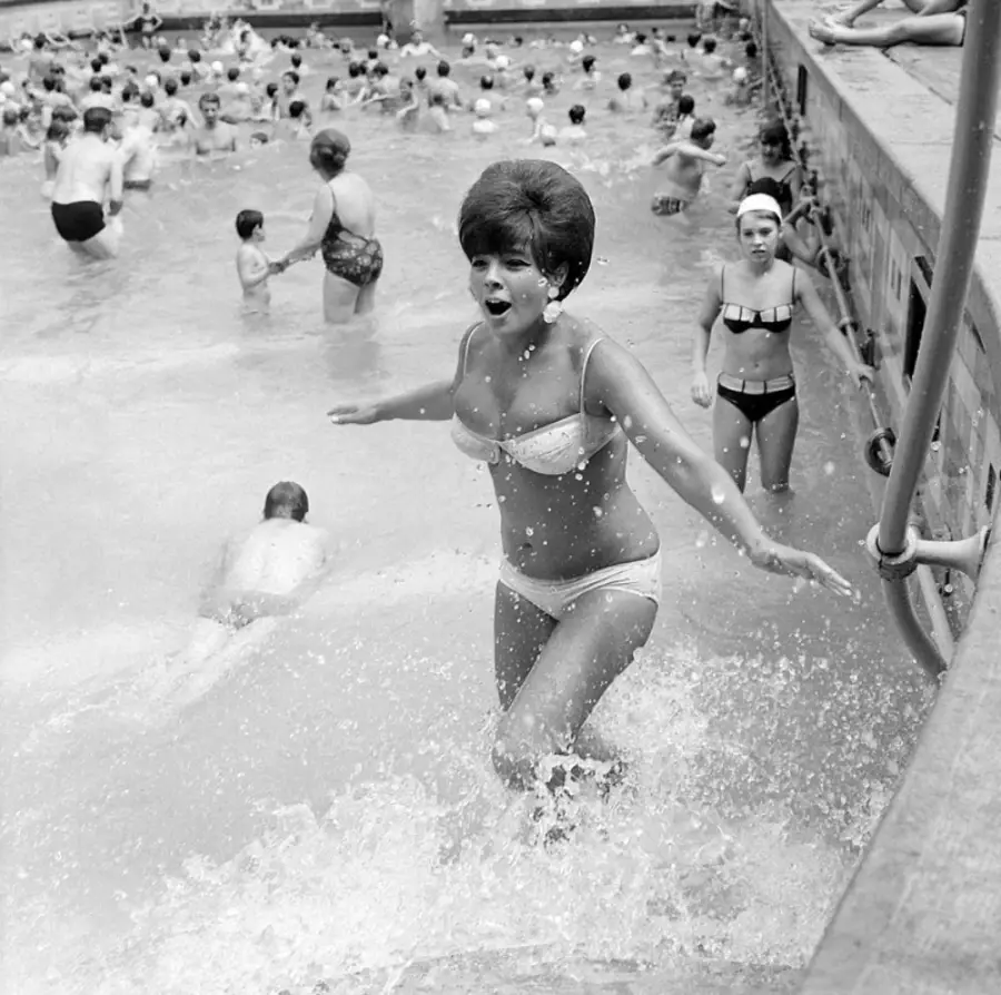 A woman in a white bikini excitedly emerges from a crowded swimming pool, splashing water. Other swimmers, adults and children, are visible in the background enjoying the pool. The image is in black and white.