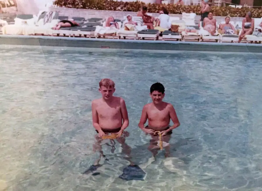 Two boys stand in a swimming pool holding goggles, while people lounge on chairs in the background near the poolside.