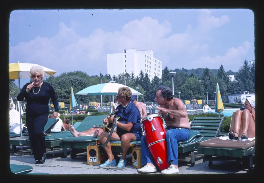 A woman sings by a pool while two shirtless men play guitar and congas; people relax on lounge chairs under umbrellas in the background.