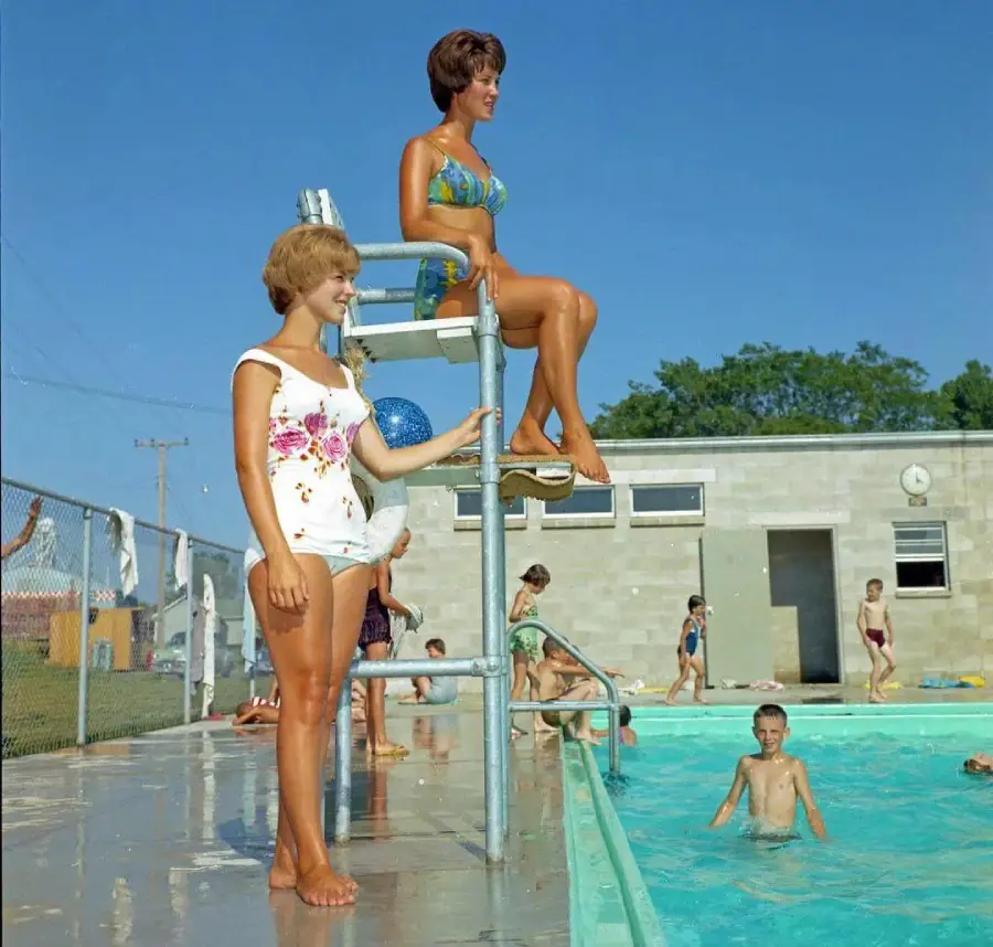 Two women in vintage swimsuits, one standing and one sitting on a lifeguard chair by a busy outdoor pool, with children swimming and playing. A building and clear blue sky are in the background.