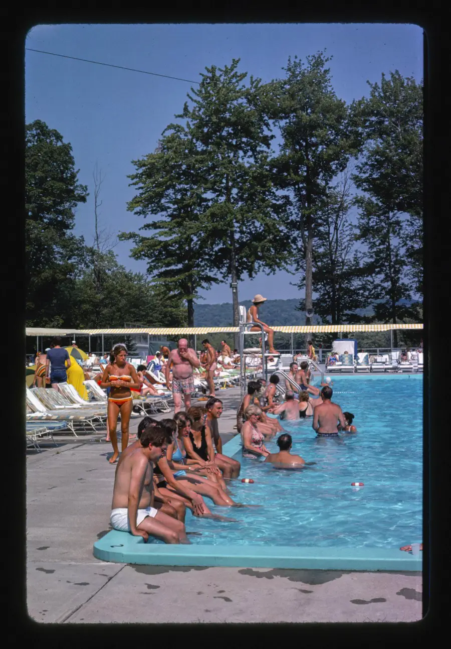 People relax by an outdoor swimming pool on a sunny day; some are in the water, others sit at the pool edge. A lifeguard watches from a high chair. Tall trees and lounge chairs are in the background.