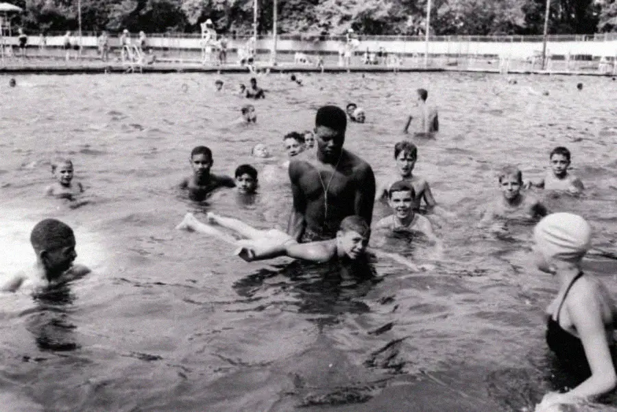 A group of children and a man are swimming and playing together in an outdoor pool. The mood is joyful, with many of the children smiling and interacting in the water.