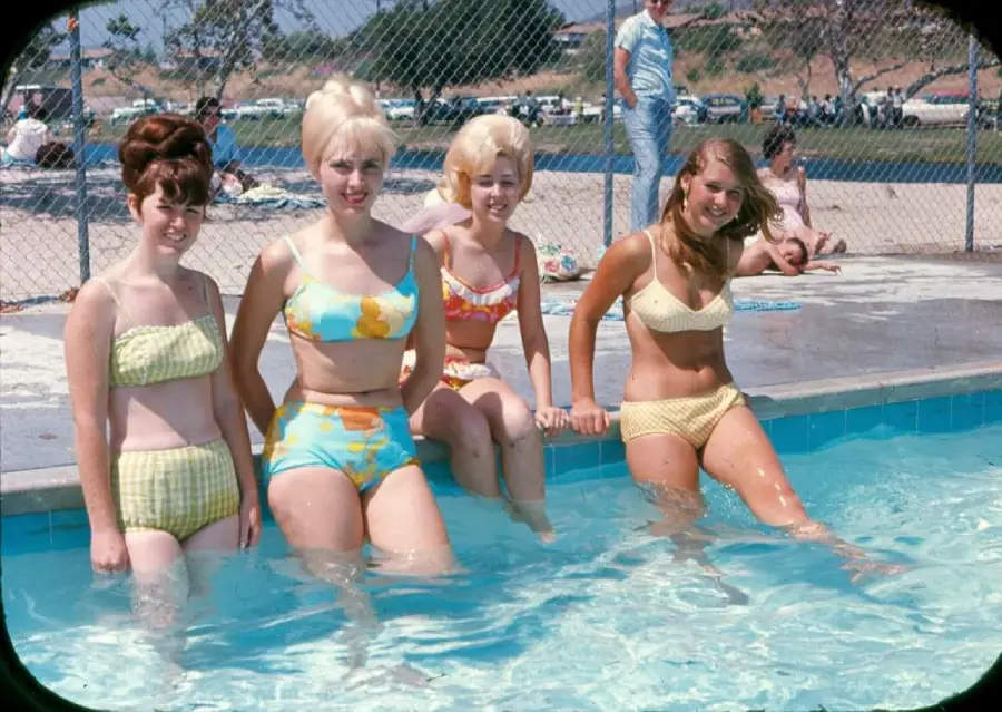Four young women in colorful vintage bikinis sit on the edge of a pool with their feet in the water, smiling. There are people in the background by the pool and cars parked behind a chain-link fence.