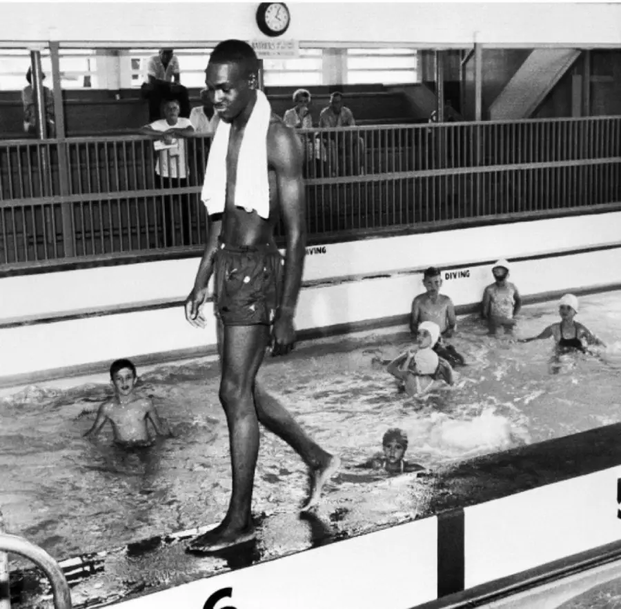 A man with a towel around his neck walks by an indoor pool as several children swim and play behind him. Spectators watch through a window above the pool. A clock is visible on the wall.