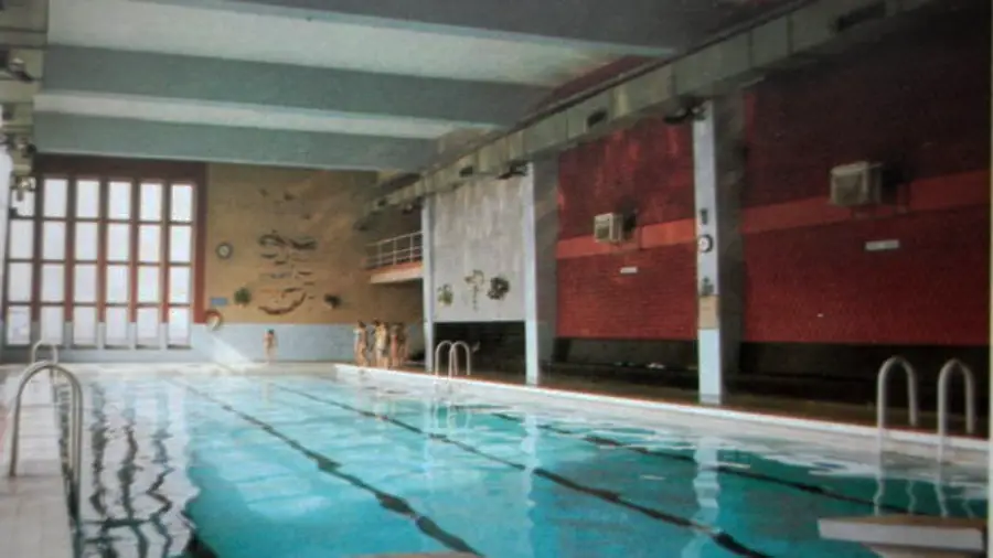 Indoor swimming pool with clear water, pool ladders, and a group of people standing near the far wall. Large windows let in daylight, and red brick walls line the sides.