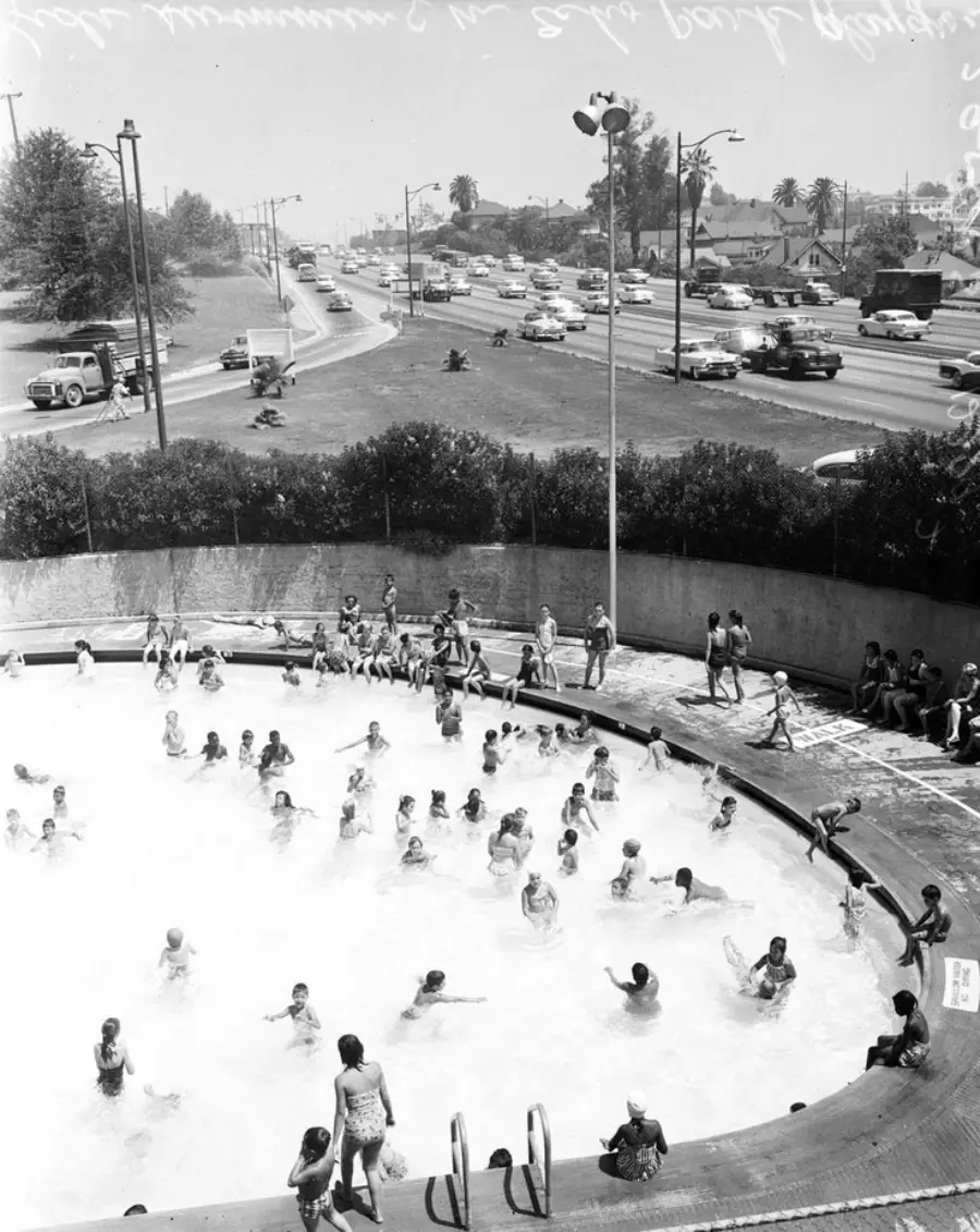 Black-and-white photo of many children swimming in a round outdoor pool, with adults watching nearby. Cars drive on a busy road and palm trees line the street in the background.