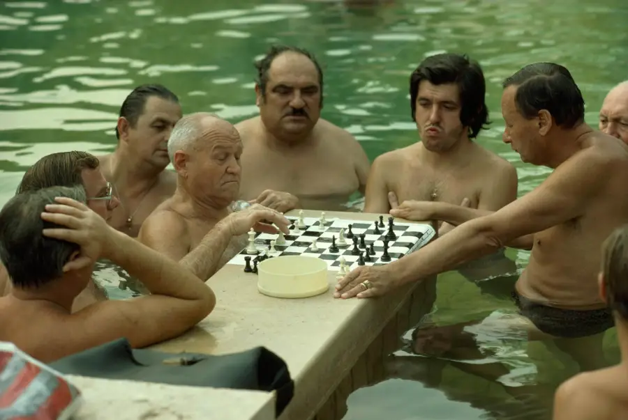 Seven men stand waist-deep in a pool, intensely focused on a game of chess played on a board balanced on the pool edge. A small round container sits next to the chessboard.