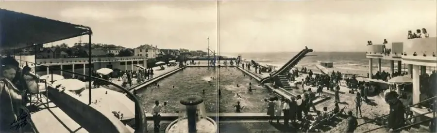 Vintage panoramic photo of a crowded seaside swimming pool with people swimming, sunbathing, and gathered on terraces; the ocean is visible in the background.