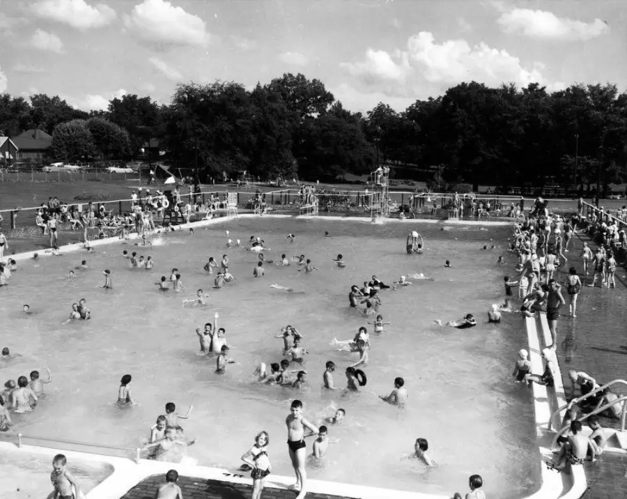 Black and white photo of a crowded outdoor swimming pool with children and adults playing in the water and others standing at the pool edge. Trees and park grounds are visible in the background under a partly cloudy sky.