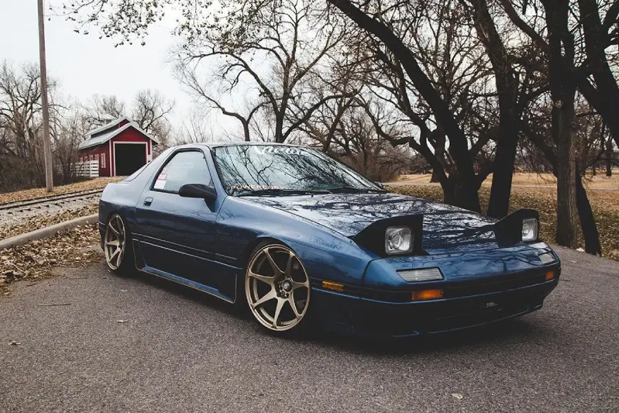 A blue sports car with pop-up headlights is parked on a road near trees and a small red building. The car sits low with large gold wheels, and fallen leaves are scattered on the ground.