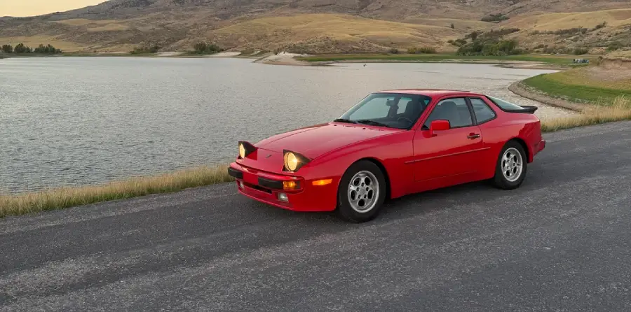 A red sports car with pop-up headlights is parked on a paved road beside a lake, with grassy hills and mountains in the background under a cloudy sky.