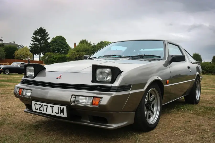 A silver classic Mitsubishi sports car with pop-up headlights is parked on grass, with trees and another car in the background under a cloudy sky. The license plate reads “C217 T JM.”