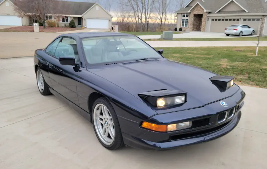 A dark blue BMW sports car with pop-up headlights is parked in a suburban driveway, with houses, grass, and leafless trees in the background.
