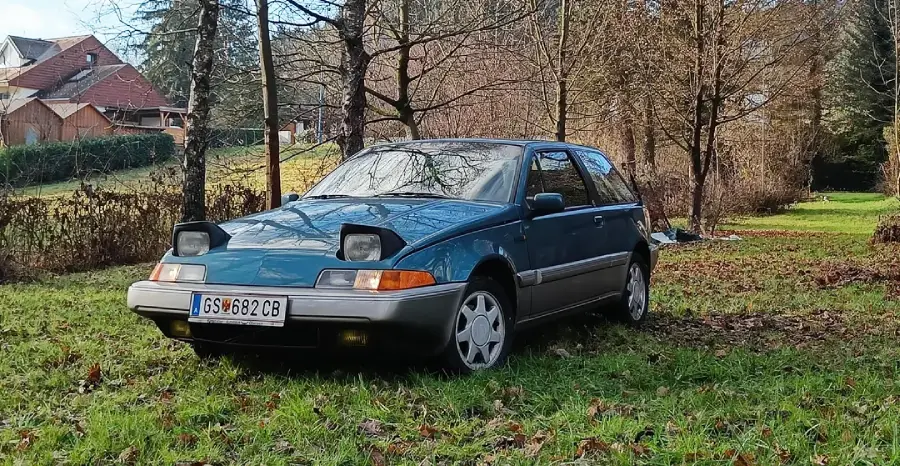 A blue vintage hatchback car with pop-up headlights is parked on a grassy area, surrounded by trees and houses in the background.