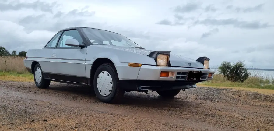 A silver two-door sports car with pop-up headlights is parked on a gravel road next to grass, under a cloudy sky near a body of water.