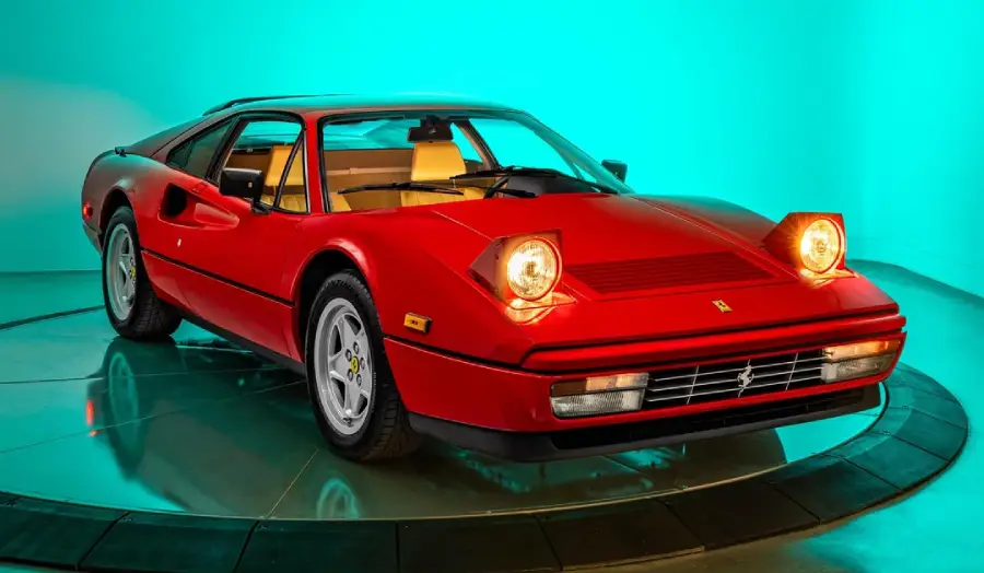 A classic red Ferrari sports car with pop-up headlights illuminated, displayed on a rotating platform against a teal-lit background.