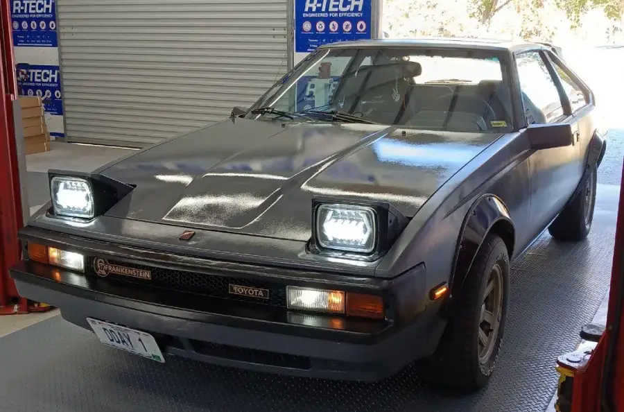 A black 1980s Toyota sports car with pop-up headlights is parked indoors near a garage door. The headlights are turned on, and the car appears to be in a workshop or service area.