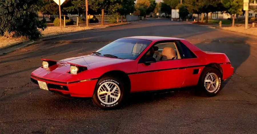 A red two-door sports car with pop-up headlights is parked on a quiet residential street at sunset. Trees and houses are visible in the background.