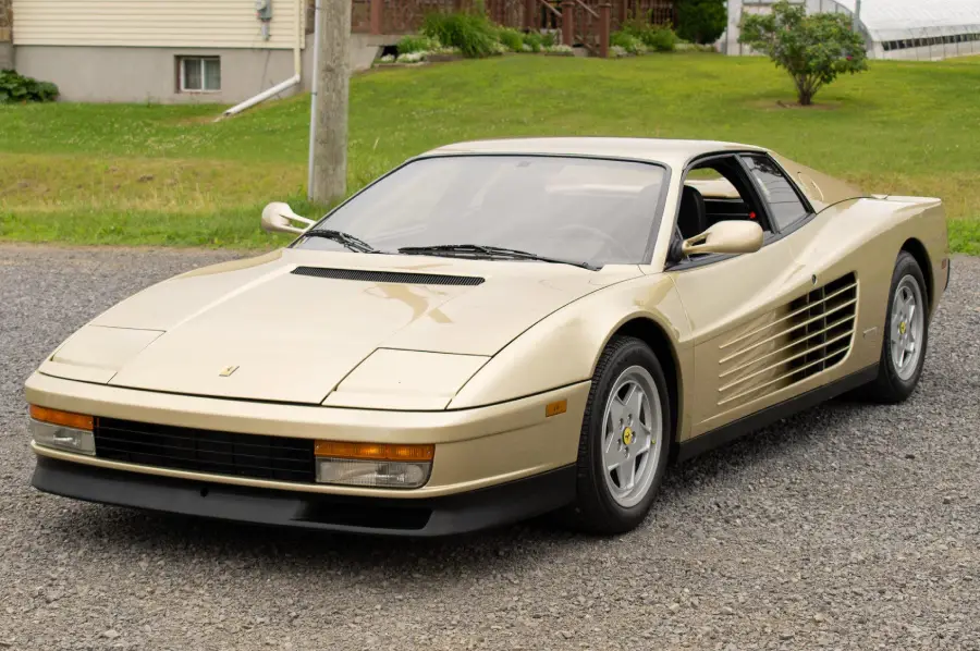 A cream-colored Ferrari Testarossa is parked on a gravel driveway, with grass and houses visible in the background.