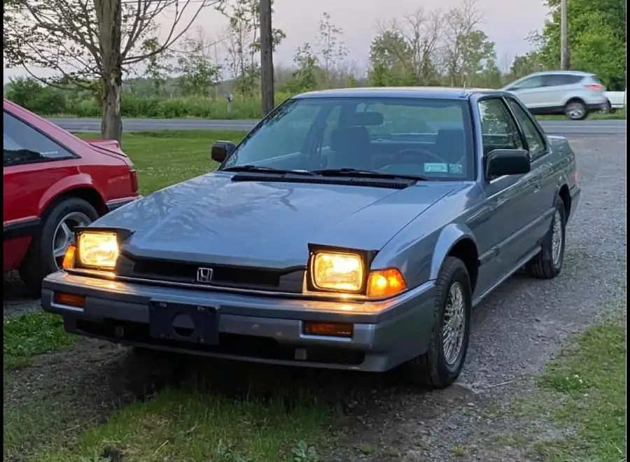A blue 1980s Honda Accord coupe with headlights on is parked on gravel next to grass. Another red car is partially visible on the left, and a road with a passing SUV is in the background.