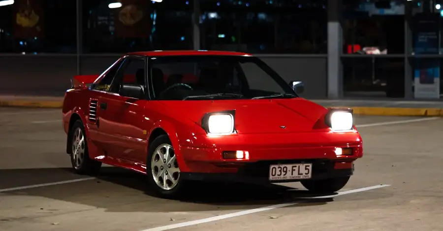 A red classic sports car with pop-up headlights on, parked alone in a well-lit lot at night.