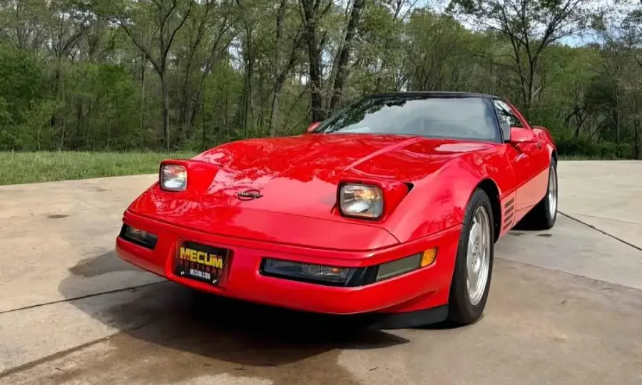 A red sports car with pop-up headlights is parked on a driveway surrounded by greenery and trees. The car has a sleek, aerodynamic design and a “Mecum” license plate on the front.