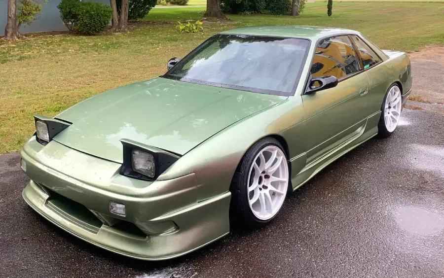 A metallic green Nissan 240SX coupe with pop-up headlights, white aftermarket wheels, and lowered suspension is parked on a wet pavement near grass and trees.