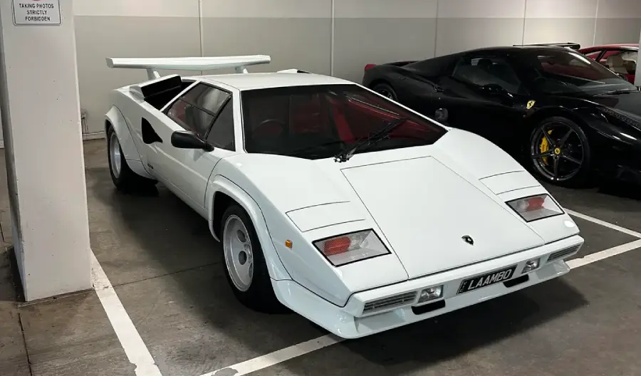 A white Lamborghini Countach is parked indoors next to a black sports car. The car features sharp angles, pop-up headlights, and a large rear wing.