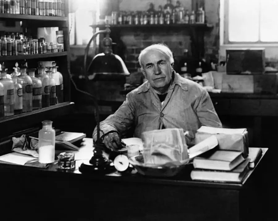 An older man sits at a cluttered desk in a laboratory, surrounded by bottles, books, and scientific equipment, with shelves and a window in the background.