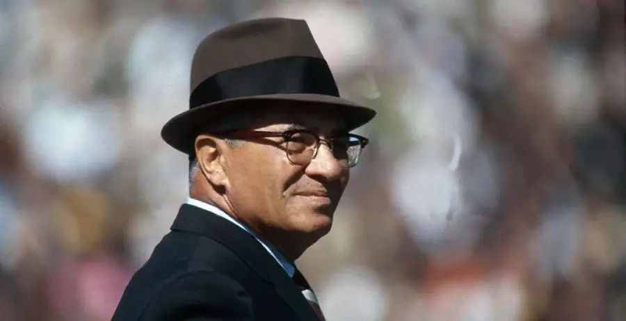 An older man in a suit, tie, and brown fedora hat stands outdoors, smiling slightly. The background is blurred, suggesting a crowd or event.