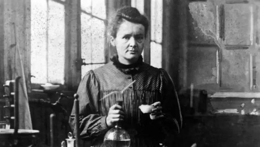 A woman in old-fashioned clothing stands in a laboratory, surrounded by scientific equipment and glassware, holding a small container in one hand and looking toward the camera.