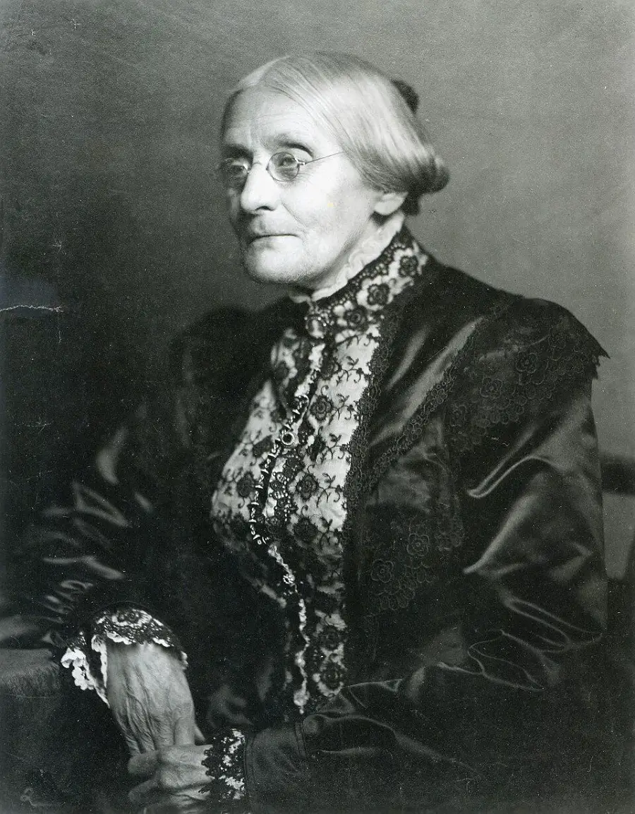 An older woman with glasses and hair in a bun, wearing a high-collared, lace-trimmed dark dress, sits facing slightly to the side in a black-and-white portrait.