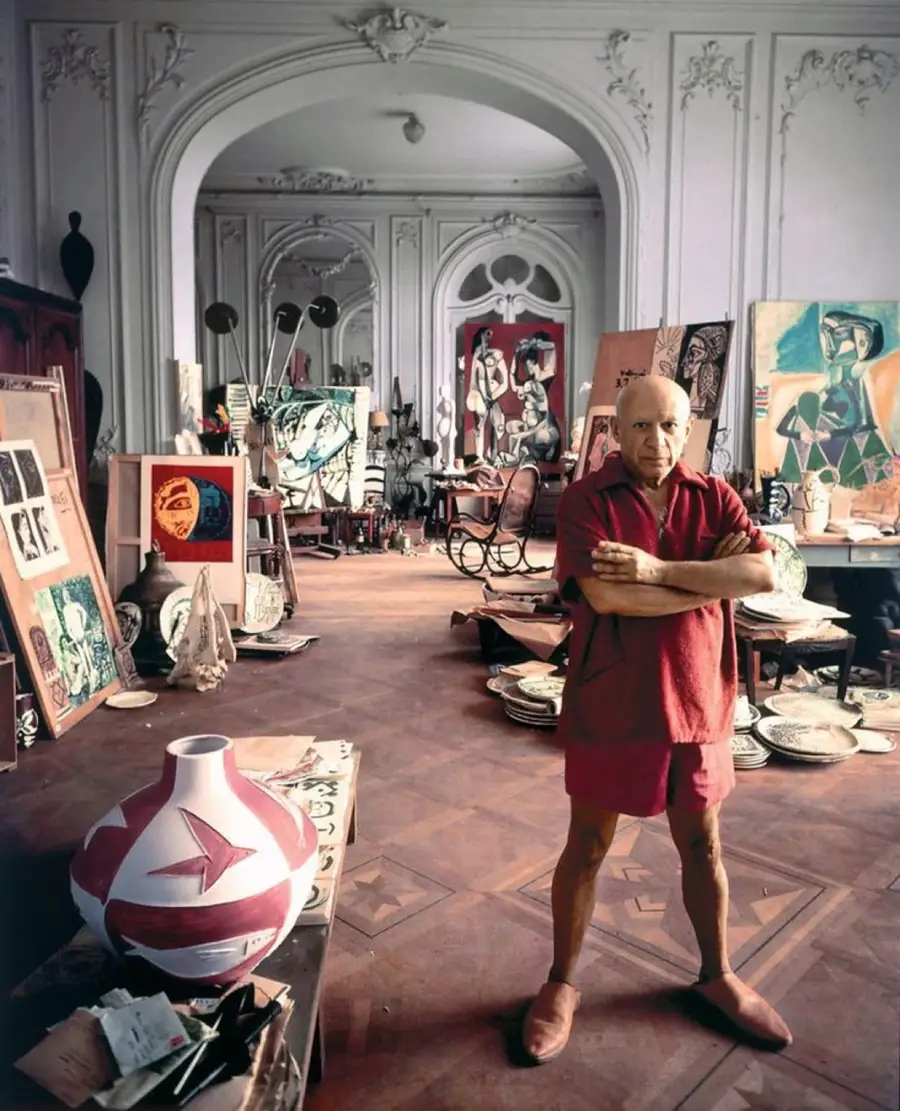 An older man in a red shirt and shorts stands with arms crossed in a spacious, ornate studio filled with paintings, sculptures, and pottery scattered around the room.