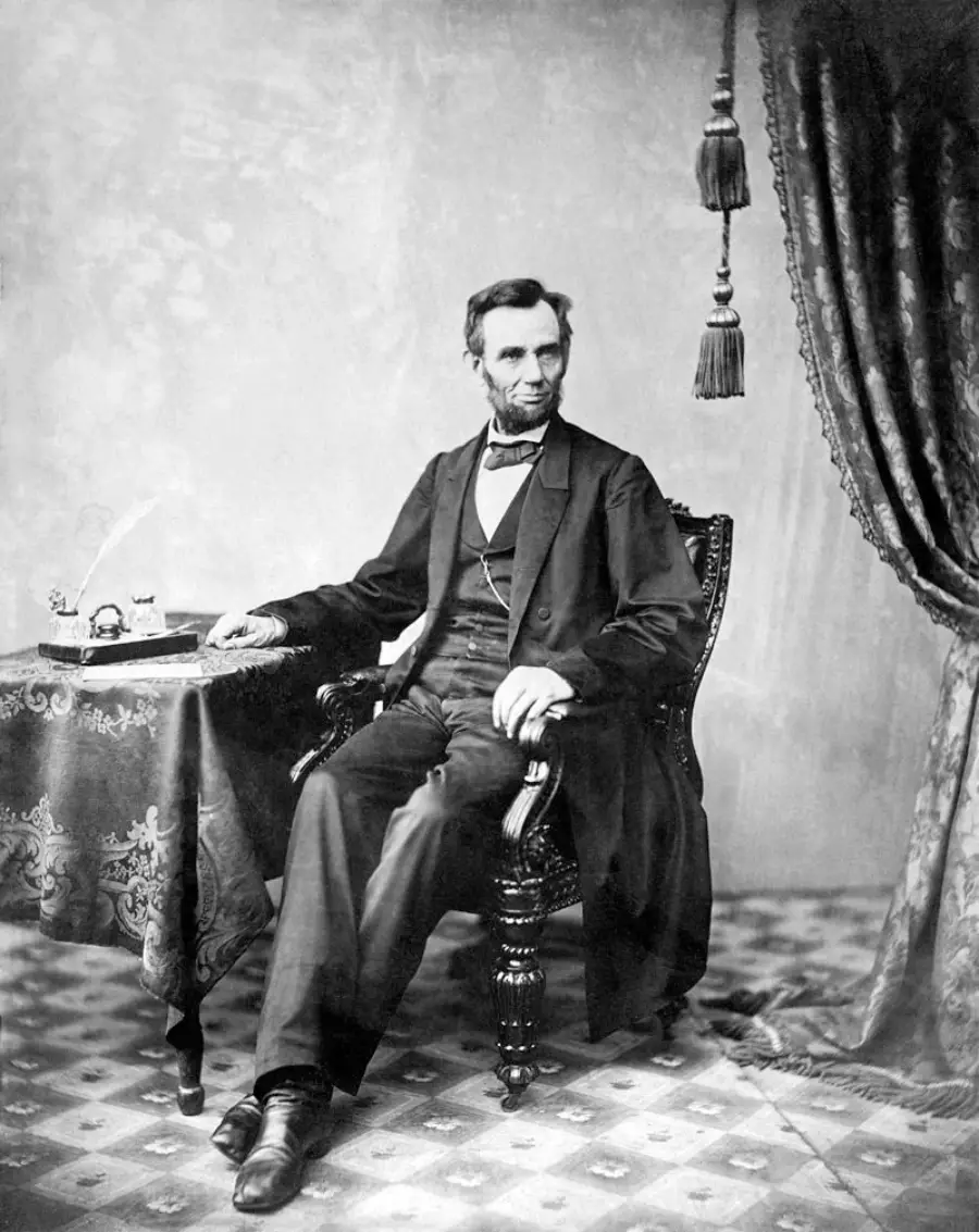 A man in a suit sits in an ornate chair beside a table with papers and a quill, against a patterned backdrop with a curtain and tassels. The photo is black and white and has a vintage feel.