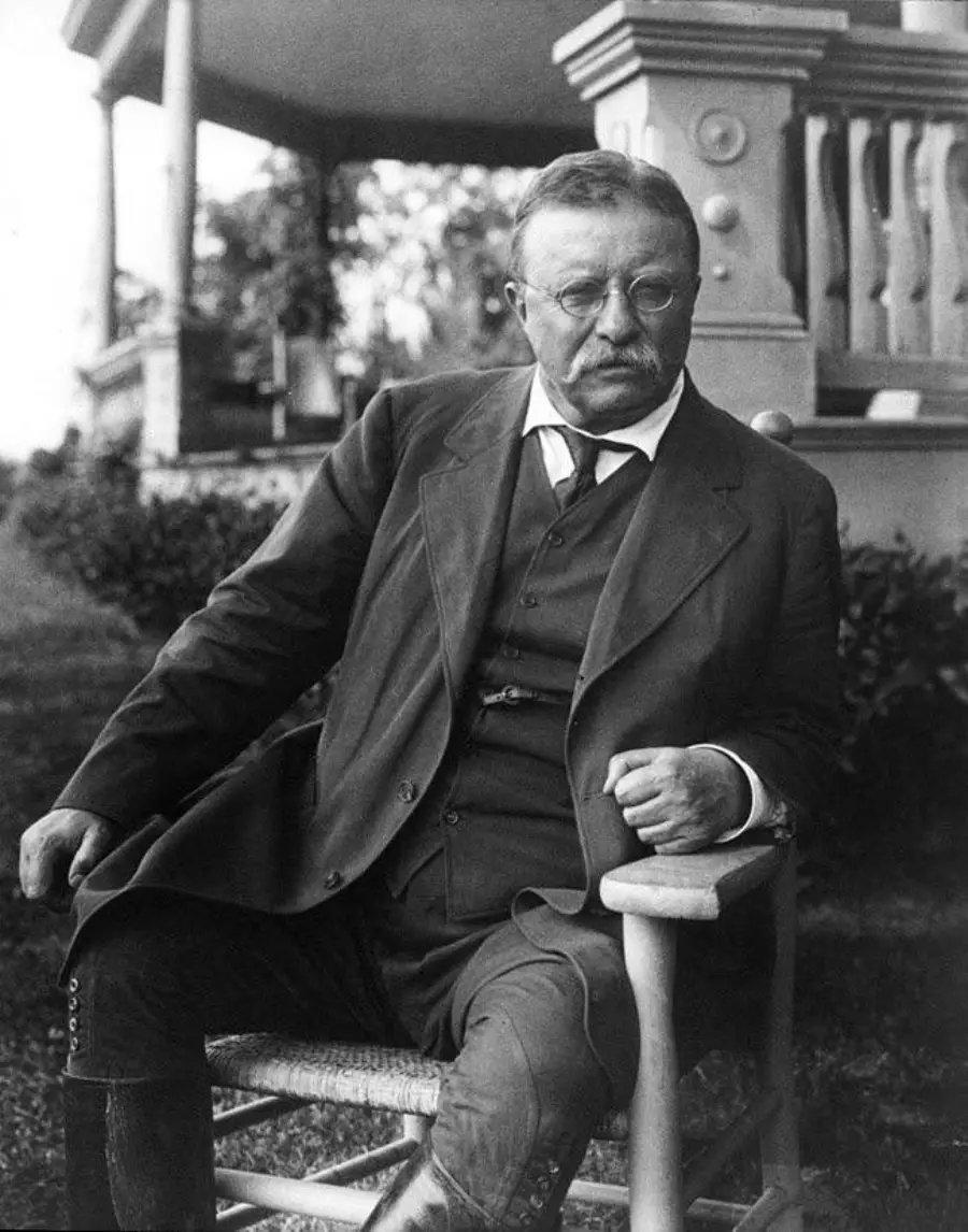 A man with glasses and a mustache sits in a wooden chair outside, dressed in a suit with a vest and tie. A porch with columns and railings is visible in the background.