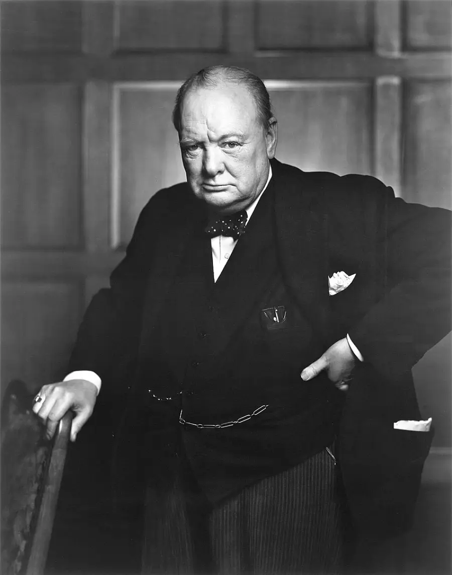 An older man in a dark suit and bow tie stands with one hand on his hip and the other on a chair, looking forward with a serious expression, in front of a wood-paneled wall. Black and white photo.