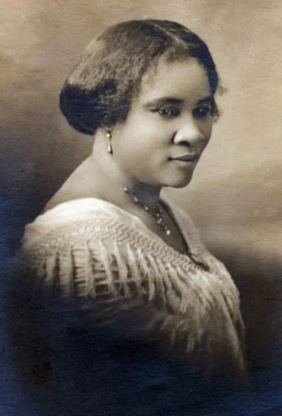 A sepia-toned portrait of a woman with styled hair, wearing earrings, a necklace, and a textured white dress, looking to the side and slightly smiling against a plain background.