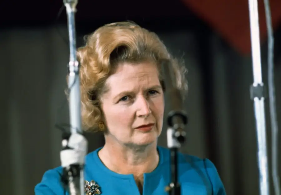 A woman with blonde, curled hair, wearing a blue jacket and a brooch, stands between two microphones, looking forward with a serious expression.