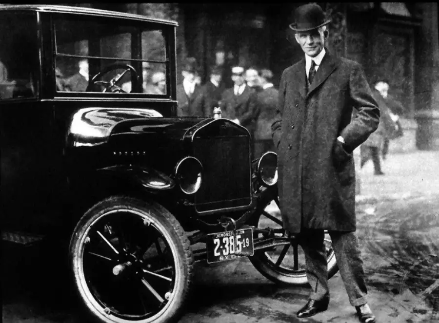 A man in a long coat and bowler hat stands smiling next to a vintage car on a city street, with several people and buildings in the background.