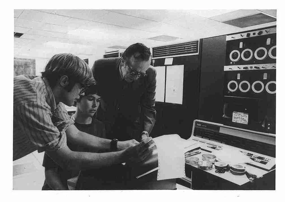 Three people examine documents in front of a vintage computer system with tape reels and control panels in an office setting, possibly engaging in collaborative work or troubleshooting.