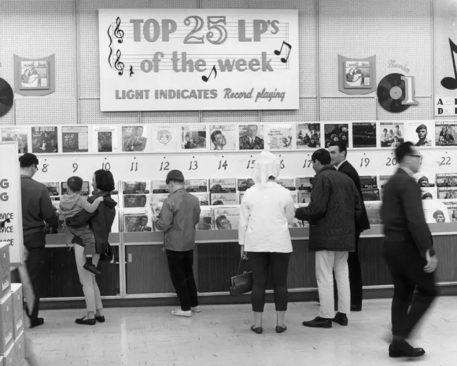 Shoppers browse records beneath a “Top 25 LP’s of the week” sign in a vintage music store. Albums are displayed on shelves, each numbered. People of various ages are looking at the record selection.