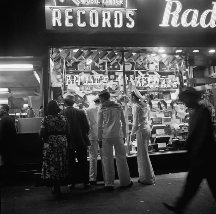 Three sailors in uniform and two civilians stand outside a brightly lit music store window at night, looking at records and radios on display. Neon signs above the storefront read “Records” and “Radios.”
