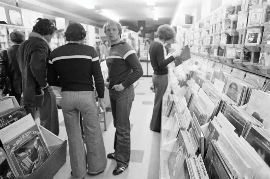 Black-and-white photo of people browsing vinyl records in a store. Three men are talking near display bins, while another person looks at records on a shelf. The store appears busy and well-stocked.